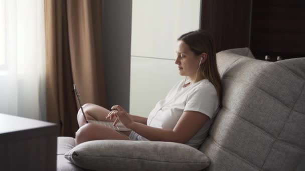 白いTシャツの女性はヘッドフォンの窓の近くのアームチェアに座っており、早朝だけで自宅で隔離されたノートブックで動作します。コンピューター上のキーボードにテキストを入力する — ストック動画