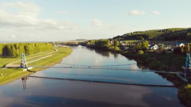 Luftaufnahme der Fußgängerbrücke über den Fluss, Südural, Russland. Per Drohne über einen breiten Fluss, an dessen Ufern dichte grüne Vegetation liegt. Die Sonne scheint in den blauen Himmel und Wolken ziehen auf — Stockvideo
