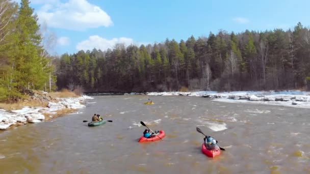 Luftaufnahme einer Gruppe von Männern, die auf hellen Schlauchbooten mit Rudern in den Händen durch die raue Strömung eines dunklen Flusses treiben. Am Ufer liegt Schnee, es gibt grüne Bäume — Stockvideo