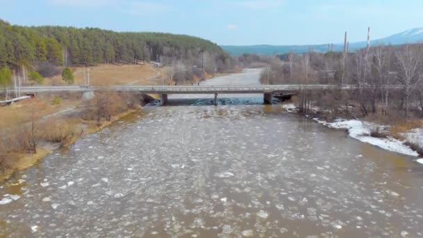 Luftaufnahme des Eisverlaufs auf dem Fluss. Die Menschen stehen auf der Brücke über den Fluss und beobachten, wie das Eis den Fluss hinunter treibt. Eis ist in viele Stücke gebrochen und der stürmische Fluss trägt es — Stockvideo