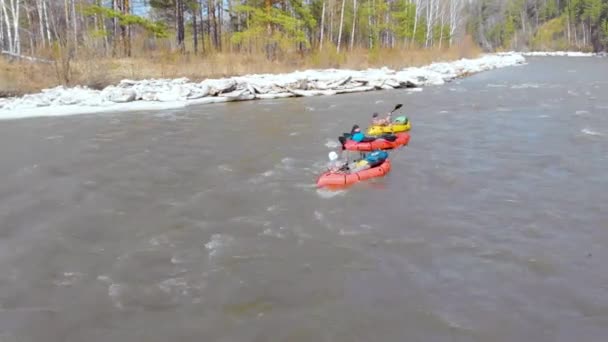 Aerial view of group of men who are floating on bright inflatable boats with oars in their hands down the rough current of a dark river. On the banks there is snow, there are green trees — Stock Video