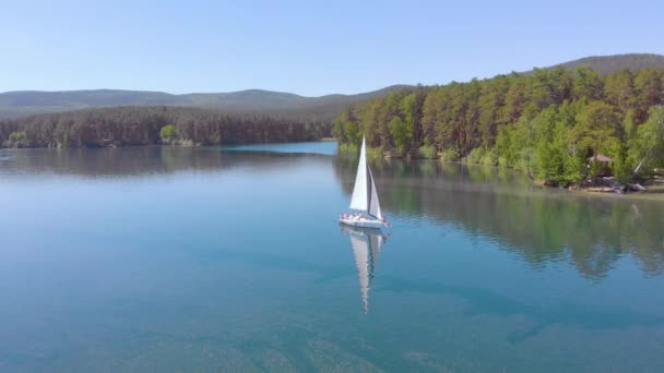 Vista aérea do barco à vela andando sozinho em um amplo lago azul claro em uma manhã ensolarada. Belo céu azul claro. Vegetação densa verde nas margens. Atmosfera pacífica. A navegar. Uma vida luxuosa — Vídeo de Stock