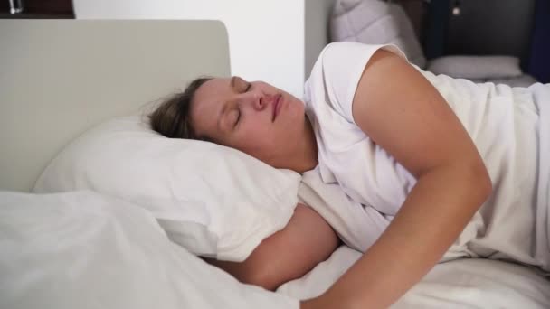 若い疲れの女性は、悪い騒々しい隣人に悩まされ、枕で彼女の耳をカバーし、スリープ状態にしようとします。不眠症に苦しむ。朝のベッドの下のカバーの下の枕の上に白いパジャマで眠る — ストック動画