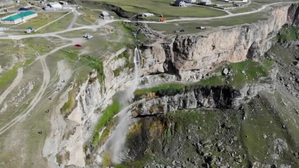 Vista aérea de una cascada alta en Daguestán. El agua corre por el acantilado a la luz del sol. La meseta de piedra está cubierta de vegetación y carreteras donde los coches conducen. Reflejos de arco iris del agua — Vídeos de Stock