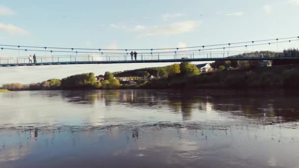 Luchtfoto van de voetgangersbrug over de rivier, Zuid-Oeral, Rusland. Vliegen op drone over brede rivier op de oevers waarvan dichte groene vegetatie. De zon schijnt aan de blauwe hemel en wolken bewegen. — Stockvideo
