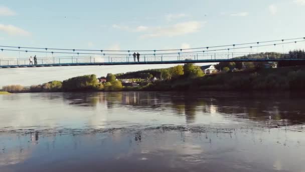 Luftaufnahme der Fußgängerbrücke über den Fluss, Südural, Russland. Per Drohne über einen breiten Fluss, an dessen Ufern dichte grüne Vegetation liegt. Die Sonne scheint in den blauen Himmel und Wolken ziehen auf — Stockvideo