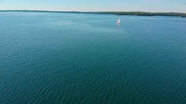 Vista aérea do barco à vela andando sozinho em um amplo lago azul claro em uma manhã ensolarada. Belo céu azul claro. Vegetação densa verde nas margens. Atmosfera pacífica. A navegar. Uma vida luxuosa — Vídeo de Stock