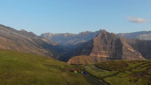 Vista aérea de los turistas que se despiertan y se encuentran con el amanecer en la roca en Daguestán. Coche rojo y tiendas de campaña contra el telón de fondo de las altas montañas. Valle de la montaña y vegetación en la cresta de piedra del Cáucaso — Vídeos de Stock