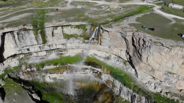 Flygfoto över ett högt vattenfall i Dagestan. Vatten rusar ner för klippan i solljus. Stenplatån är täckt med grönska och vägar där bilar kör. Regnbågsreflektioner från vatten — Stockvideo
