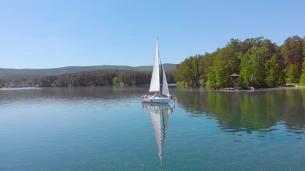 Vista aérea del velero caminando solo en un amplio lago azul claro en una mañana soleada. Hermoso cielo azul claro. Vegetación verde densa en las orillas. Ambiente pacífico. Navegando. Una vida de lujo — Vídeos de Stock