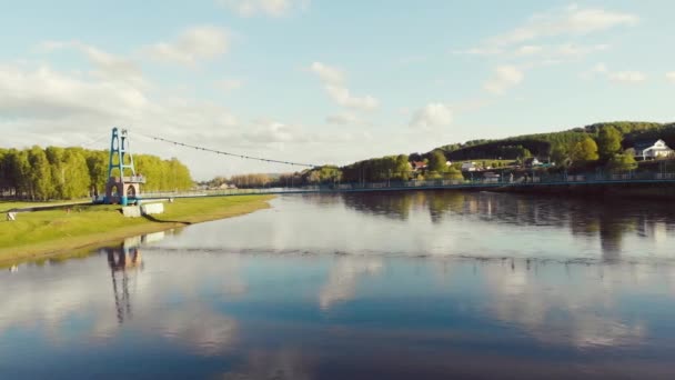 Nehrin üzerindeki yaya köprüsünün havadan görünüşü, Güney Ural, Rusya. Geniş nehrin üzerinde, yoğun yeşil bitki örtüsünün bulunduğu kıyılarda insansız hava aracıyla uçuyorlar. Güneş mavi gökyüzünde parlıyor ve bulutlar hareket ediyor. — Stok video