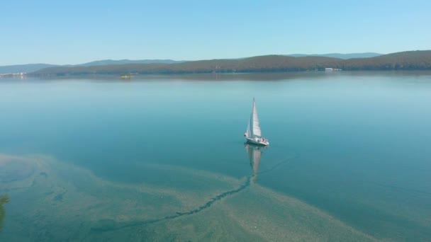 Vista aérea do barco à vela andando sozinho em um amplo lago azul claro em uma manhã ensolarada. Belo céu azul claro. Vegetação densa verde nas margens. Atmosfera pacífica. A navegar. Uma vida luxuosa — Vídeo de Stock