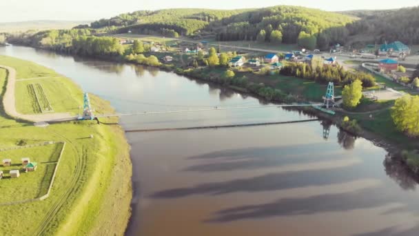 Flybilde av gangbroen over elven, Sør-Ural, Russland. Flyr på drone over bred elv på breddene av den tette grønne vegetasjonen. Sola skinner på den blå himmelen, og skyene beveger seg – stockvideo