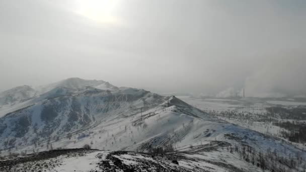 Vista aérea da fábrica. As colinas de inverno estão cobertas de neve. Atrás de fios e suportes eléctricos. A fumaça venenosa acrílica vem das chaminés da planta. Poluição ambiental — Vídeo de Stock