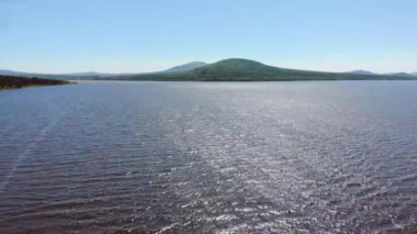 Açık mavi gölün havadan görünüşü. Koyu yemyeşil ağaçlar kıyılarda yetişir. Hafif bir esinti. Dalgalanmalar gölde yüksek gri dağların arka planına doğru akar. Güneş ışıkla oynuyor. Yaz