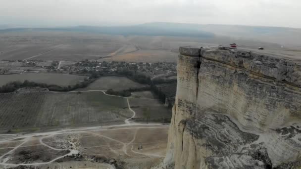 Luchtfoto van White Rock, Krim schiereiland. Er staan auto 's bovenaan de hoge klif. Toeristen wandelen over het rotsplateau. Trails lopen vanaf de bodem van de berg, velden worden groen — Stockvideo