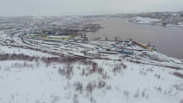 Légi felvétel a kikötőről, Murmanszk, Oroszország. Hosszú gyártócsarnokok a Barents-tenger havas partján. Anyagok hajókon történő kirakodása és berakodása. Nagy vasdaruk a parton. Sarki élet — Stock videók
