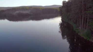 Açık mavi gölün havadan görünüşü. Koyu yemyeşil ağaçlar kıyılarda yetişir. Hafif bir esinti, sis. Dalgalanmalar gölde yüksek gri dağların arka planına doğru akar. Güneş ışıkla oynuyor. Yaz