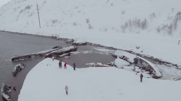 Légi felvétel a hajó temetőjéről, Teriberka, Murmanszk. A turisták a Barents-tenger homokos partján sétálnak, amelyet hó borít. Tél. Félelmetes hajócsontvázak állnak ki a vízből. — Stock videók