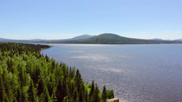 Vue aérienne d'un lac bleu clair profond. Des arbres luxuriants épais poussent sur les rives. Légère brise soufflant. Des ondulations traversent le lac sur fond de hautes montagnes grises. Le soleil joue avec la lumière. Été — Video