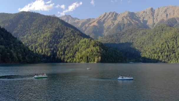 Luftaufnahme vom Ritsa-See, Abchasien. Die Menschen fahren mit Katamaranen und Booten auf der ebenen Oberfläche des türkisfarbenen Sees. Strahlend grüne Bäume bedecken die Ufer. Berge und Wolken sind in der Ferne zu sehen — Stockvideo
