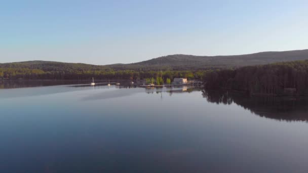 Aerial view of clear deep lake. Thick lush trees grow on the banks. Light breeze blowing. Sailboats are sailing. Ripples run across the lake against the backdrop of high gray mountains. Summer — Stock Video