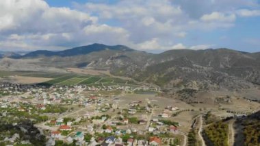 Tepenin önündeki vadinin havadan görüntüsü. Ovada renkli evler var. Tarlalar birçok yola göre meydanlara ayrılır. Gökyüzünde bir sürü bulut var.