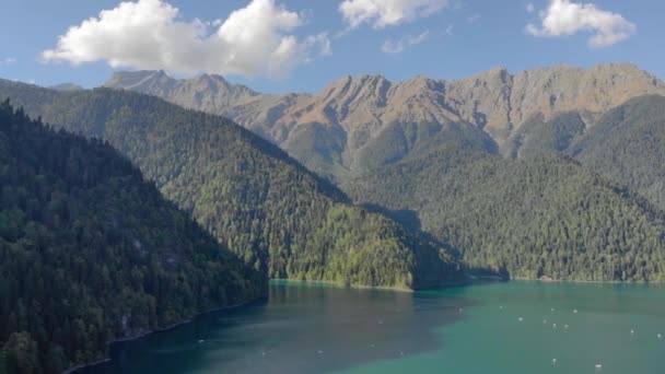 Vista aérea del lago Ritsa, Abjasia. La gente monta catamaranes y barcos en la superficie plana del lago turquesa. Los árboles verdes brillantes cubren las orillas. Montañas y nubes se pueden ver en la distancia — Vídeo de stock