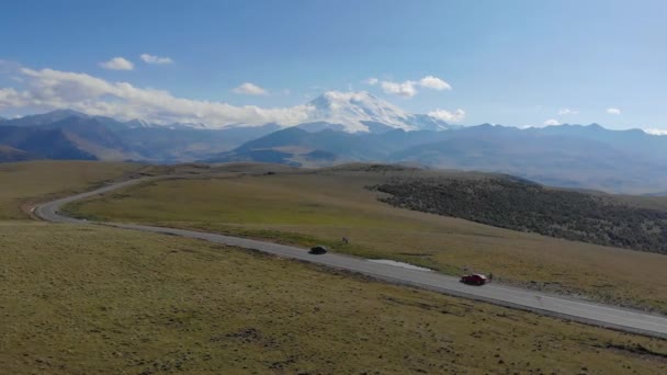 Widok z lotu ptaka na drogę biegnącą przez łańcuch górski Kaukazu, region Elbrus. Samochody stoją na drodze wśród zielonych łąk. Słońce oświetla szczyty gór. — Wideo stockowe