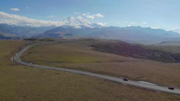 Widok z lotu ptaka na drogę biegnącą przez łańcuch górski Kaukazu, region Elbrus. Samochody stoją na drodze wśród zielonych łąk. Słońce oświetla szczyty gór. — Wideo stockowe