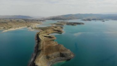 Baraj gölünün havadan görünüşü. Gök mavisi suyu olan yapay göl. Höyükler adalar gibidir. Karada kırmızı bir araba var. Bulutlar suya yansıyor. Dağlar uzaktan görülebilir.. 
