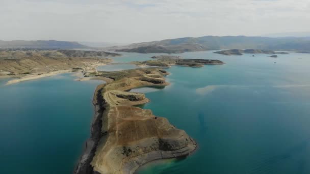 Luchtfoto van het reservoir. Kunstmatig meer met azuurblauw water. De heuvels zijn als eilanden. Er staat een rode auto op het land. De wolken worden weerspiegeld in het water. Bergen zijn zichtbaar in de verte. — Stockvideo