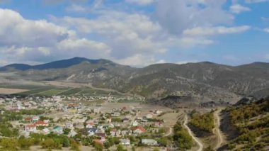 Tepenin önündeki vadinin havadan görüntüsü. Ovada renkli evler var. Tarlalar birçok yola göre meydanlara ayrılır. Gökyüzünde bir sürü bulut var.