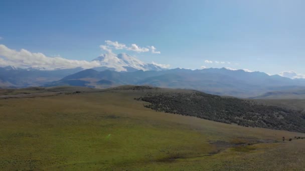从空中俯瞰穿过Elbrus地区高加索山脊的公路。汽车站在绿色草地中间的路上.游客们欣赏山景.太阳照亮了高山的最高峰 — 图库视频影像