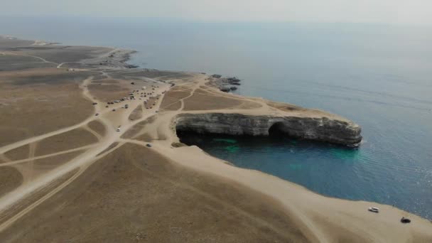 Αεροφωτογραφία του ακρωτηρίου Tarkhankut, Μαύρη Θάλασσα, χερσόνησος της Κριμαίας. Δρόμοι κατά μήκος των οποίων πηγαίνουν τα αυτοκίνητα περνούν από τη γη. Το σπήλαιο σε λευκό ασβεστολιθικό βράχο. Η θάλασσα είναι γαλάζια. Ο ήλιος φωτίζει το νερό — Αρχείο Βίντεο