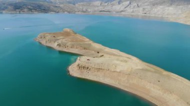 Baraj gölünün havadan görünüşü. Gök mavisi suyu olan yapay göl. Höyükler adalar gibidir. Karada kırmızı bir araba var. Bulutlar suya yansıyor. Dağlar uzaktan görülebilir.. 