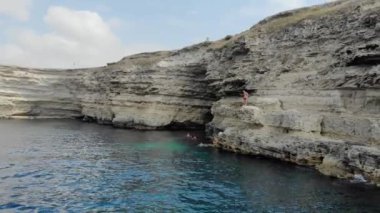 Uçurumun havadan görünen manzarası. Cape Tarkhankut, Karadeniz, Kırım yarımadası. Kırmızı şortlu adam suya atlamaya hazırlanıyor ve kayaların üzerinde duruyor. İnsanlar masmavi derin denizde yüzer.
