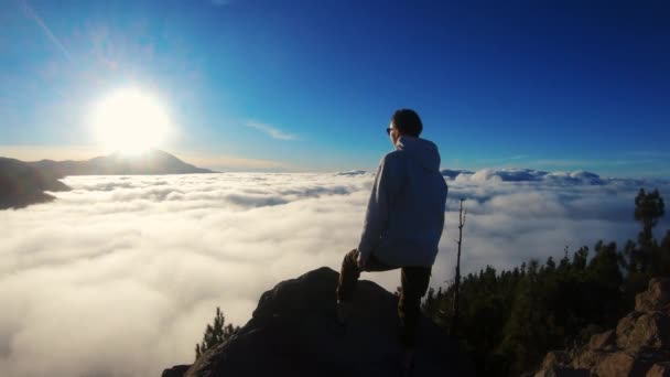 女性は山の頂上に立って、白い雲が浮かんでいる距離を見ています。深く呼吸し、幸せを感じる。青い空に山の上に輝く太陽をお楽しみください。ハイキング — ストック動画