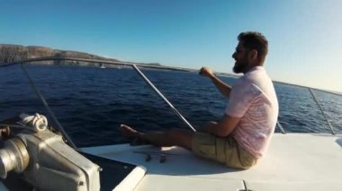 Genç adam beyaz yatla gezmekten zevk alıyor. Güvertede çıplak ayakla oturur, tırabzana tutunur ve adalara bakar. Yüzünde güneş gözlüğü var. Derin ve mutlu nefes alır.