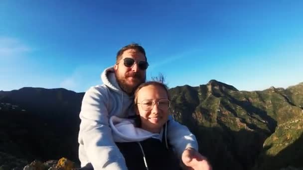 Par i kärlek klättrade till toppen och skjuta sig själva på kameran mot bakgrund av berg och havsvik. Mannen i solglasögon vinkar hand. Berg mot blå himmel och ljus sol. Jag vandrar. Smekmånad — Stockvideo