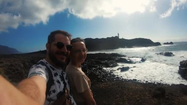 Una pareja encantadora grabándose en una cámara de video. Un hombre con gafas de sol abraza a su amada. La mujer muestra la mano en las olas. La tormenta rompe olas azules contra rocas y piedras. Senderismo. Luna de miel — Vídeo de stock