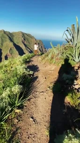 De mens loopt naar het pad op de top van de berg. De zon schijnt en de schaduw van het lichaam is zichtbaar. Benadert vrouw die stopt en foto 's maakt met smartphone. Bergen en groen zijn rondom zichtbaar — Stockvideo