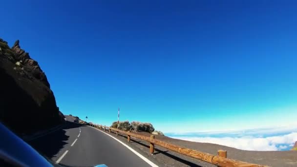 Автомобиль едет по плоской дороге вдоль скалы. Облака и холмы можно увидеть ниже. Дорога с белыми отметинами, знаками, заборами. Солнечная погода, чистое голубое небо. Стрельба со стороны синей машины — стоковое видео