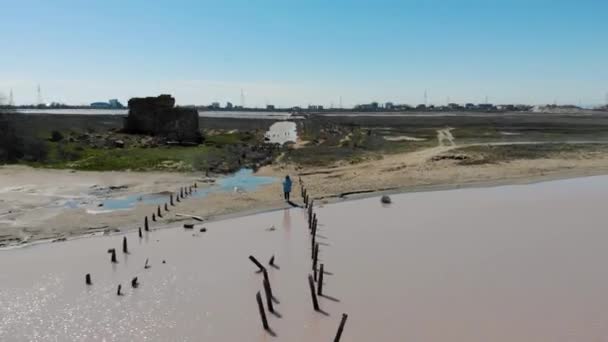 Luftaufnahme des rosa Salzsees, Halbinsel Krim. Frau in blauem Regenmantel steht am Ufer des Sees. Weg aus Holzstümpfen geht ins Wasser. See und Schwarzes Meer sind durch einen Sandstreifen getrennt. Einzigartige Natur — Stockvideo