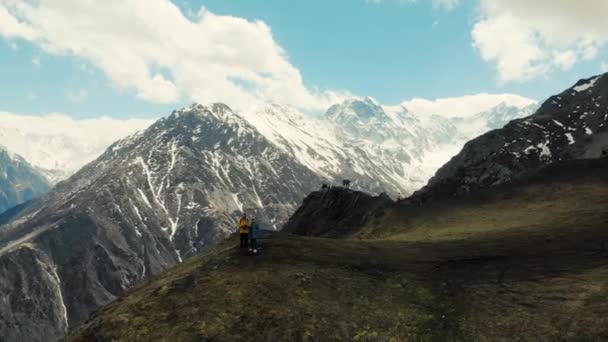 Coppia si trova sul bordo della scogliera in cima alla montagna, Fiagdon, Caucaso. Vista di enormi montagne del Caucaso. Cime di alta montagna sono coperte di neve. Le nuvole pendono dall'alto. Terribile bellezza — Video Stock