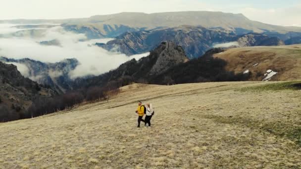 Vista aérea das montanhas do Cáucaso. Algumas pessoas estão a atravessar o campo. O céu cinzento é visível sobre os prados verdes. Nuvens brancas formam o rio. Tempo nublado — Vídeo de Stock