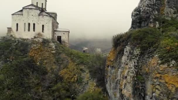 Vista aérea do templo antigo no topo da montanha. As paredes brancas da estrutura são visíveis no topo da montanha entre árvores verdes brilhantes. Nuvens grossas e nevoeiro estão descendo. Tempo nublado — Vídeo de Stock