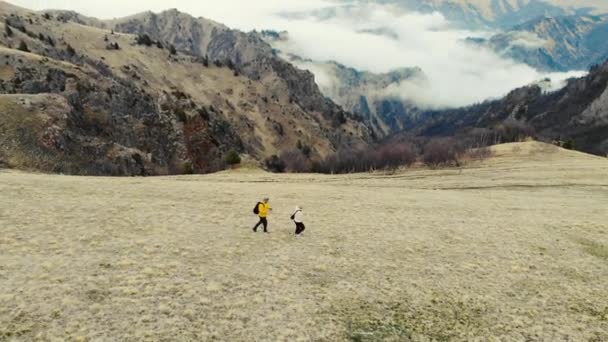Vista aérea de las montañas del Cáucaso. Un par de personas están cruzando el campo. El cielo gris es visible sobre prados verdes. Nubes blancas forman el río. Clima nublado — Vídeos de Stock