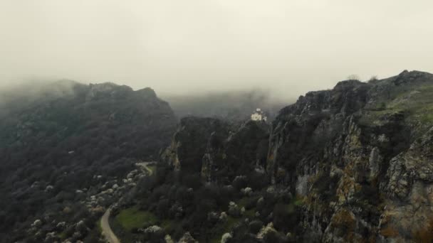 Vista aérea del antiguo templo en la cima de la montaña. Las paredes blancas de la estructura son visibles en la cima de la montaña entre árboles verdes brillantes. Nubes gruesas y niebla están bajando. Clima nublado — Vídeos de Stock