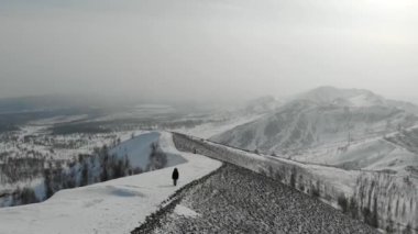 Kadının yürüdüğü dağ yolunun havadan görünüşü. Yol karla kaplı. Etrafta küçük kış tepeleri var. Ağaçlar görünür. Her yerde gri gökyüzü ve bulutlar var. Güzel kasvetli manzara